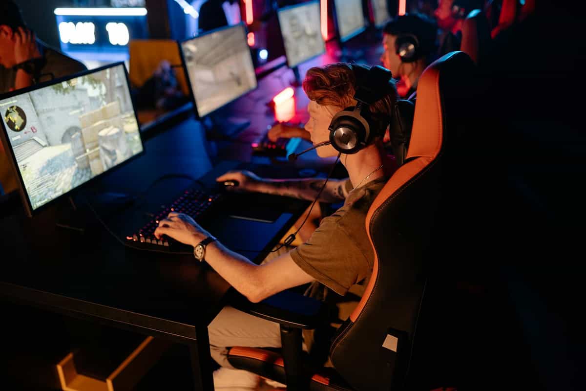 A high-angle, side view of a young man with red hair wearing a headset, intently playing a first-person shooter game in a dimly lit gaming center. Competitive CS2